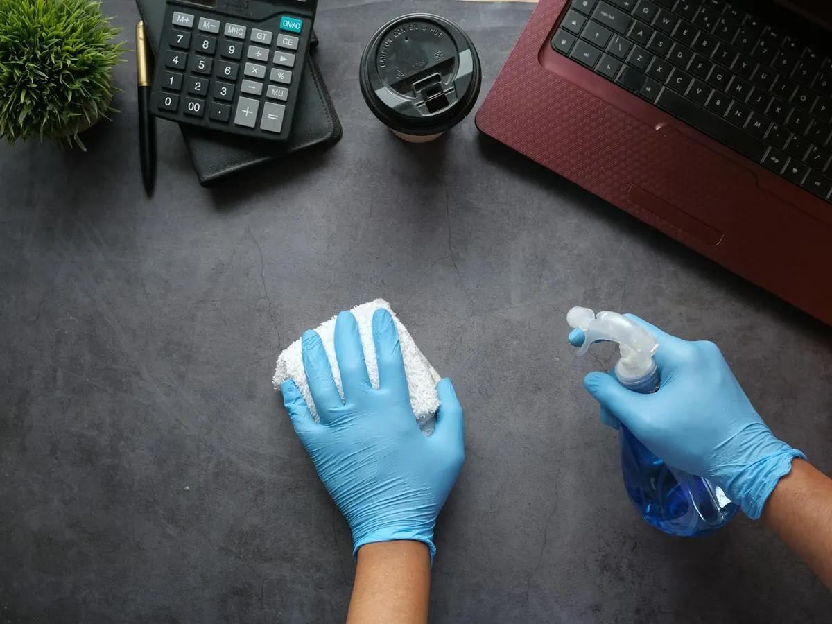 Cleaner in blue gloves wiping down a desk with spray and a cloth